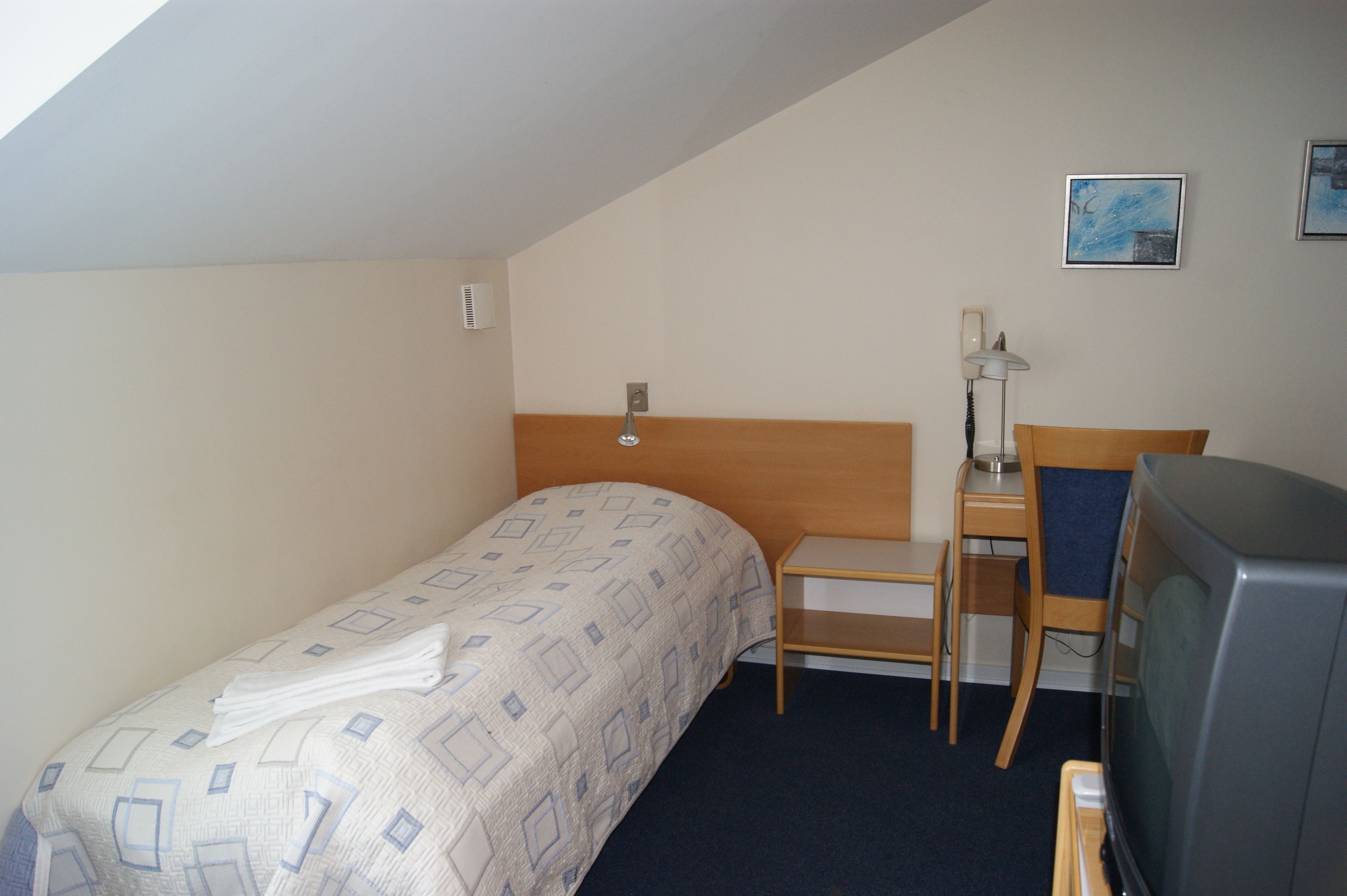 A small, simple room with a bed, a desk, a chair, a television, and a wall-mounted telephone.