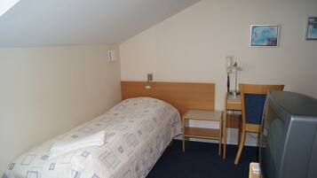 A small, simple room with a bed, a desk, a chair, a television, and a wall-mounted telephone.