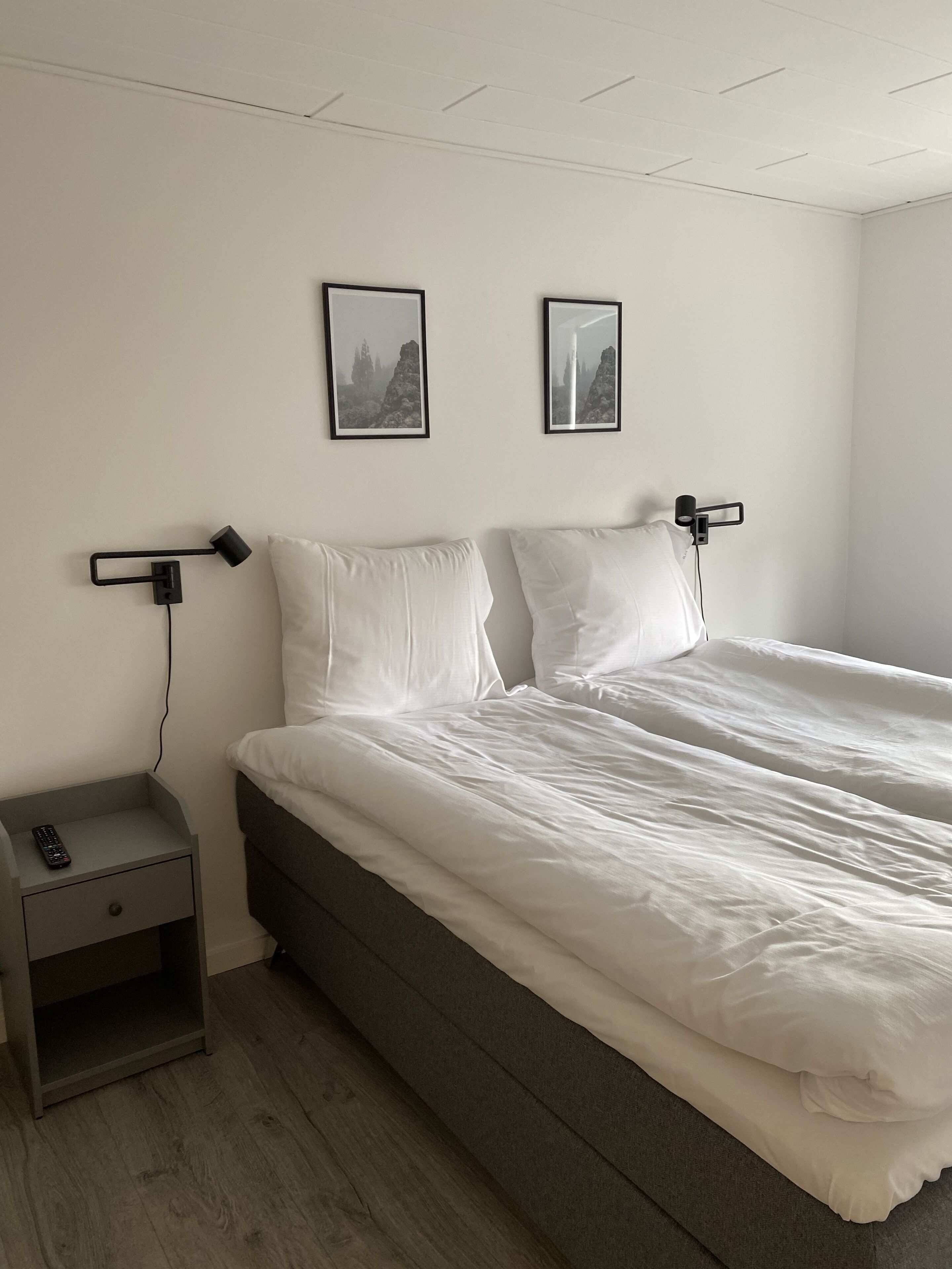 Un lit double avec une literie blanche, une table de chevet avec une télécommande et deux tableaux encadrés au mur.
