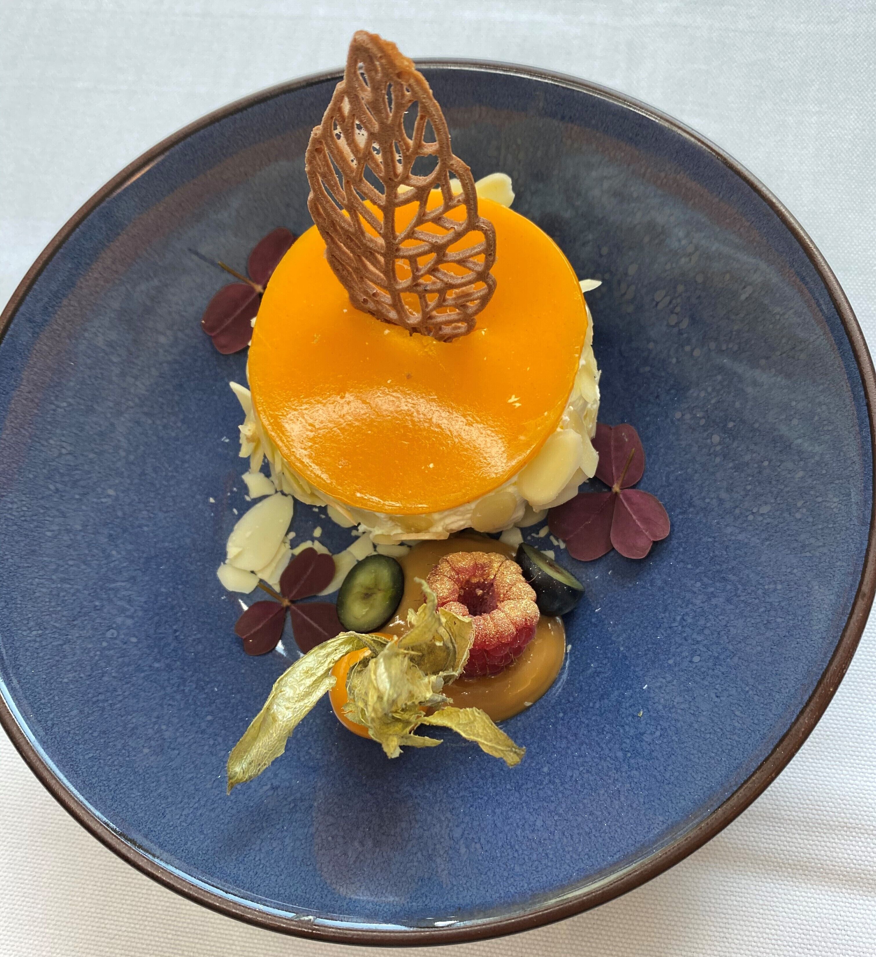 Un postre sobre un plato azul con una hoja decorativa y una fruta cubierta de azúcar.