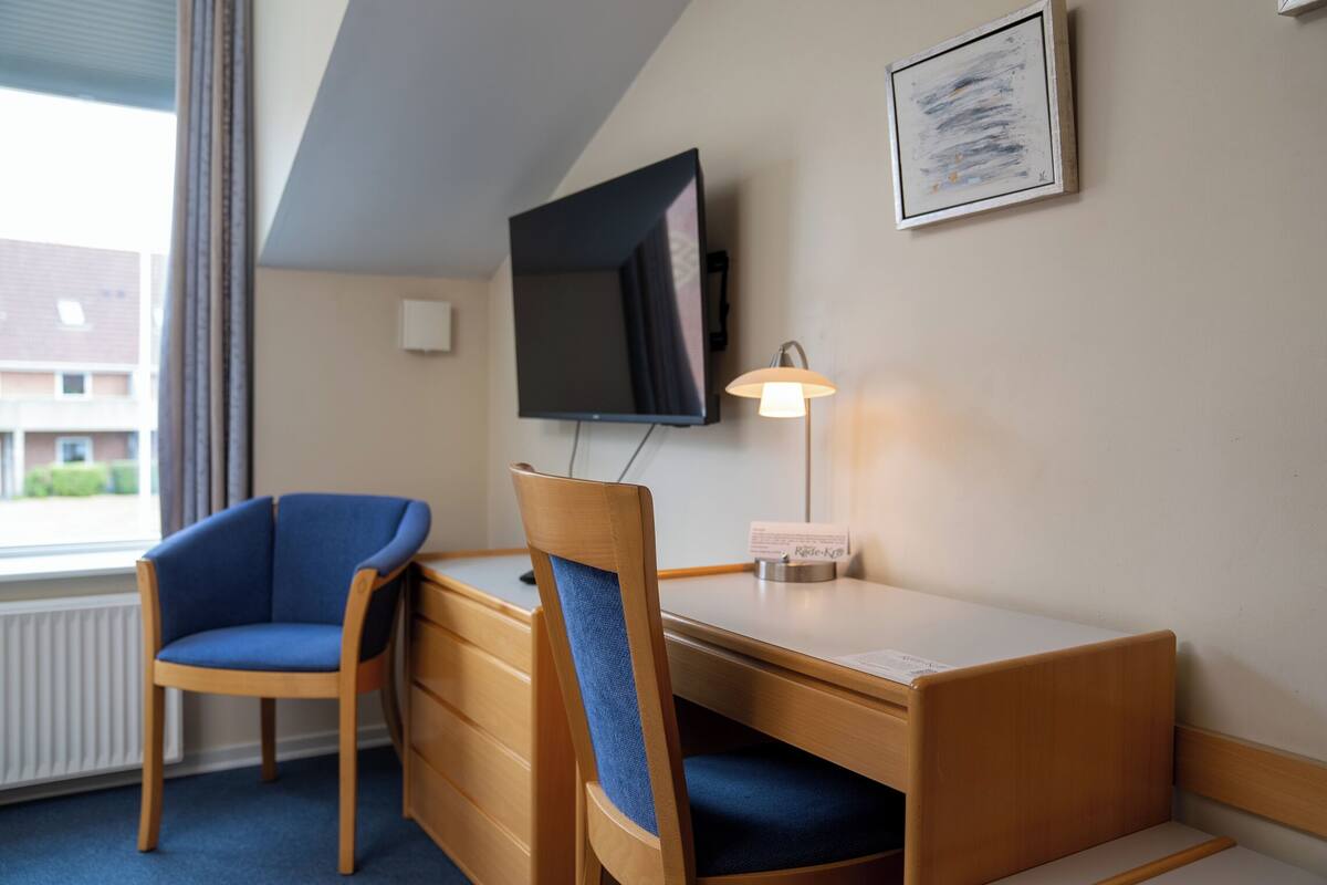 Habitación de hotel con escritorio de madera, una silla azul, televisor montado en la pared y un cuadro enmarcado.