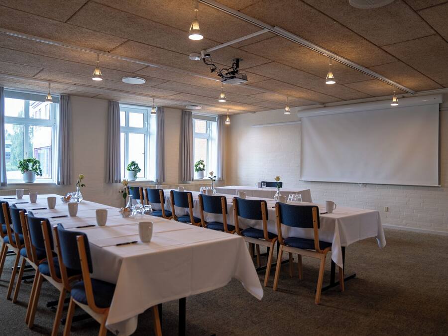 A conference room with multiple tables and chairs, a projector screen, and large windows with curtains.
