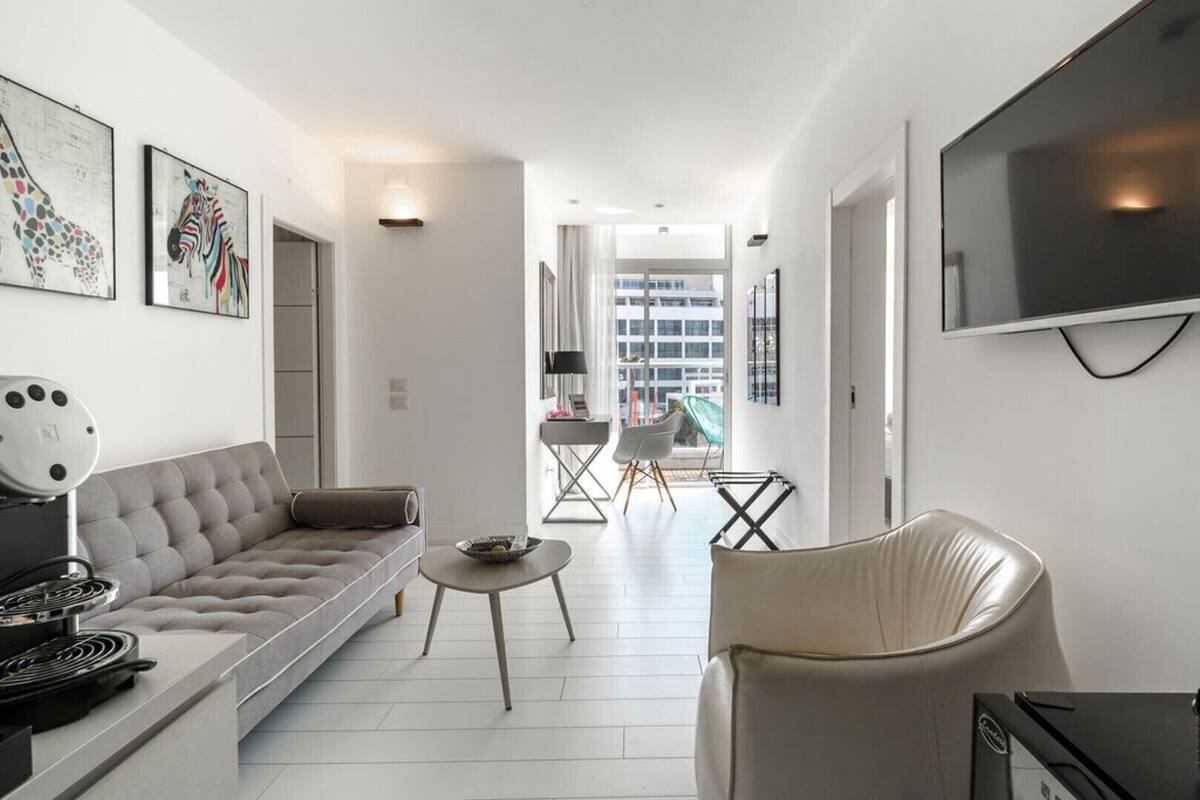 A modern living room with a sofa, a dining table, and a TV. There are framed pictures on the wall and a window with a view of buildings.
