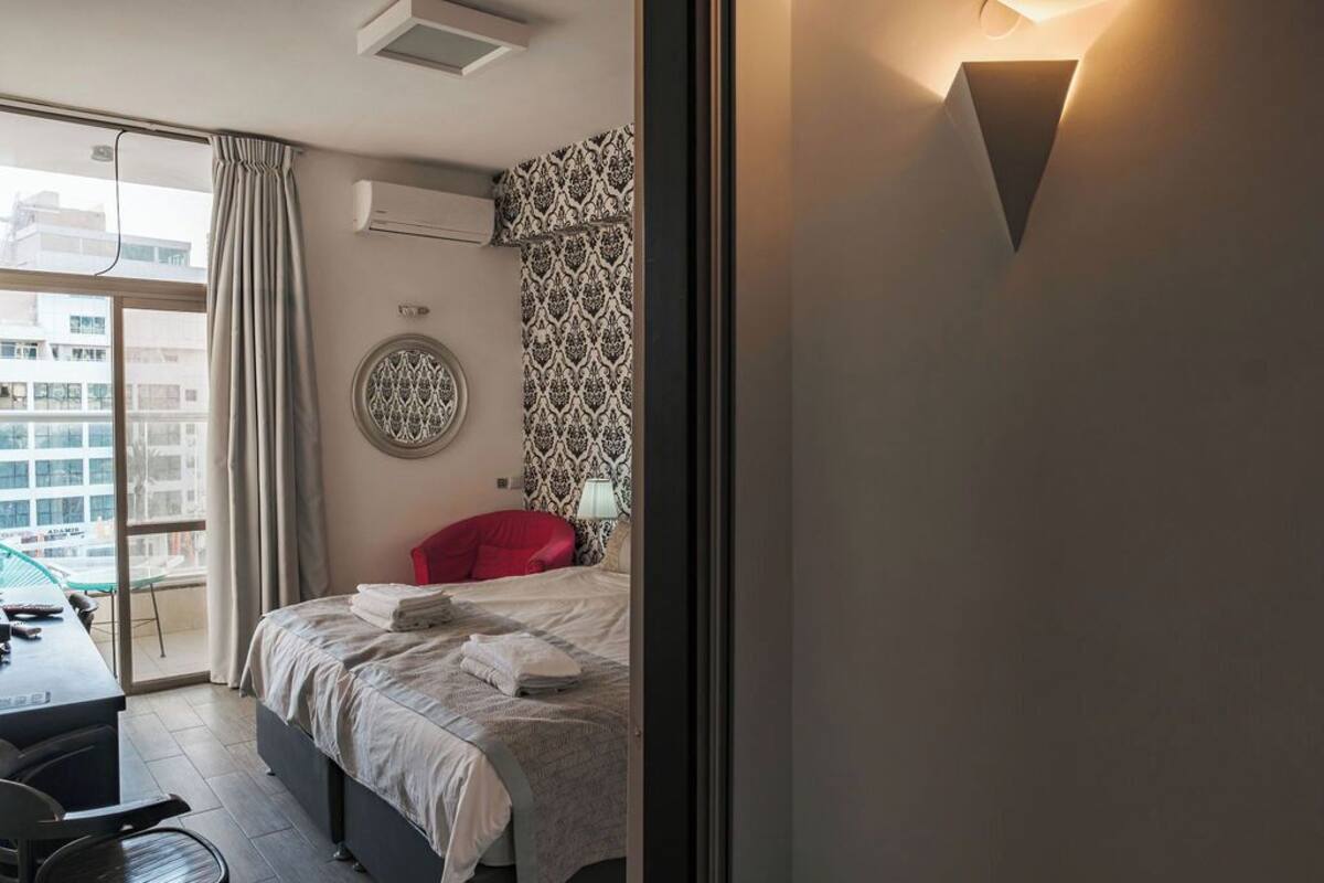 A hotel room with a bed, a desk, and a view of the city through large windows.