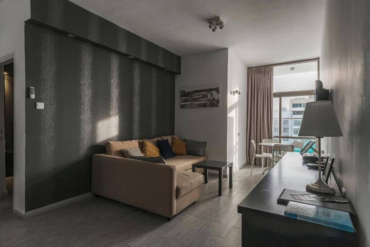 A modern living room with a sofa, coffee table, and a desk with a lamp. There is a window with curtains and a view of buildings outside.
