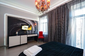 A modern hotel room with a black bed, a red chair, a desk with a computer, and a large window with curtains.