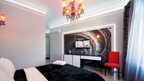 Quarto de hotel moderno com uma cama grande, uma escrivaninha com televisão, uma cadeira vermelha e um candelabro.