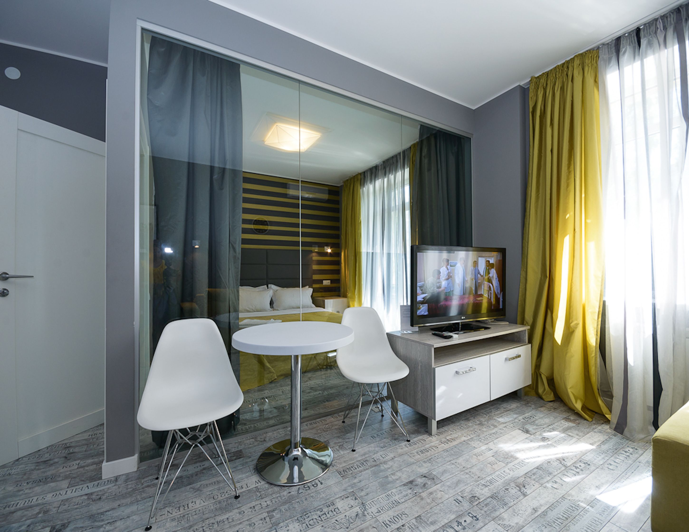 Moderni hotellihuone, jossa on sänky, televisio, ruokapöytä tuoleineen ja lasiseinä.