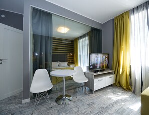 A modern hotel room with a bed, a TV, a dining table with chairs, and a glass partition.