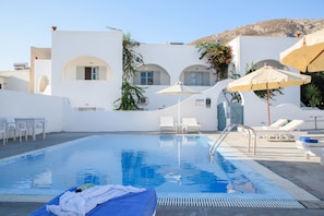 En swimmingpool med liggestole og parasoller, omgivet af hvide bygninger med buede vinduer.
