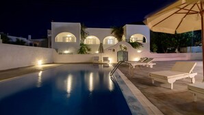 A swimming pool with lounge chairs and umbrellas, surrounded by a modern building with arched windows and a well-lit exterior. - Atlas Boutique Hotel (Santorini)