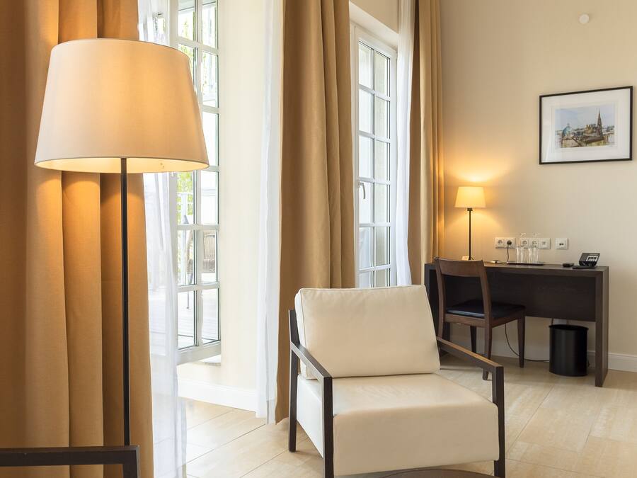 Habitación de hotel con sofá, una mesa redonda, un escritorio con silla, una lámpara y una ventana con cortinas.