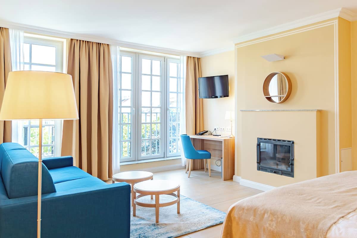 A hotel room with a sofa, desk, chair, TV, fireplace, and large windows.