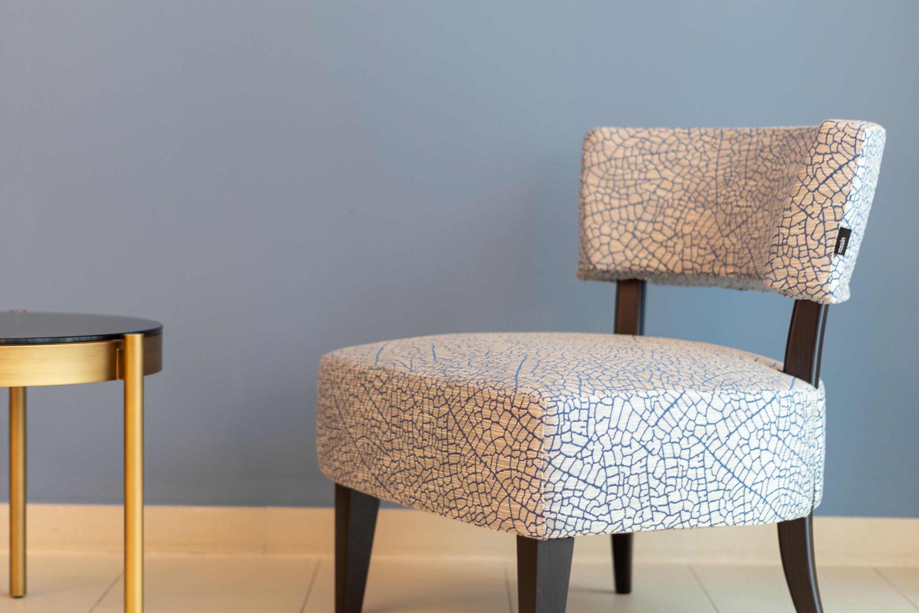 A chair with a cracked pattern upholstery, a round table with a gold frame, and a plain wall.