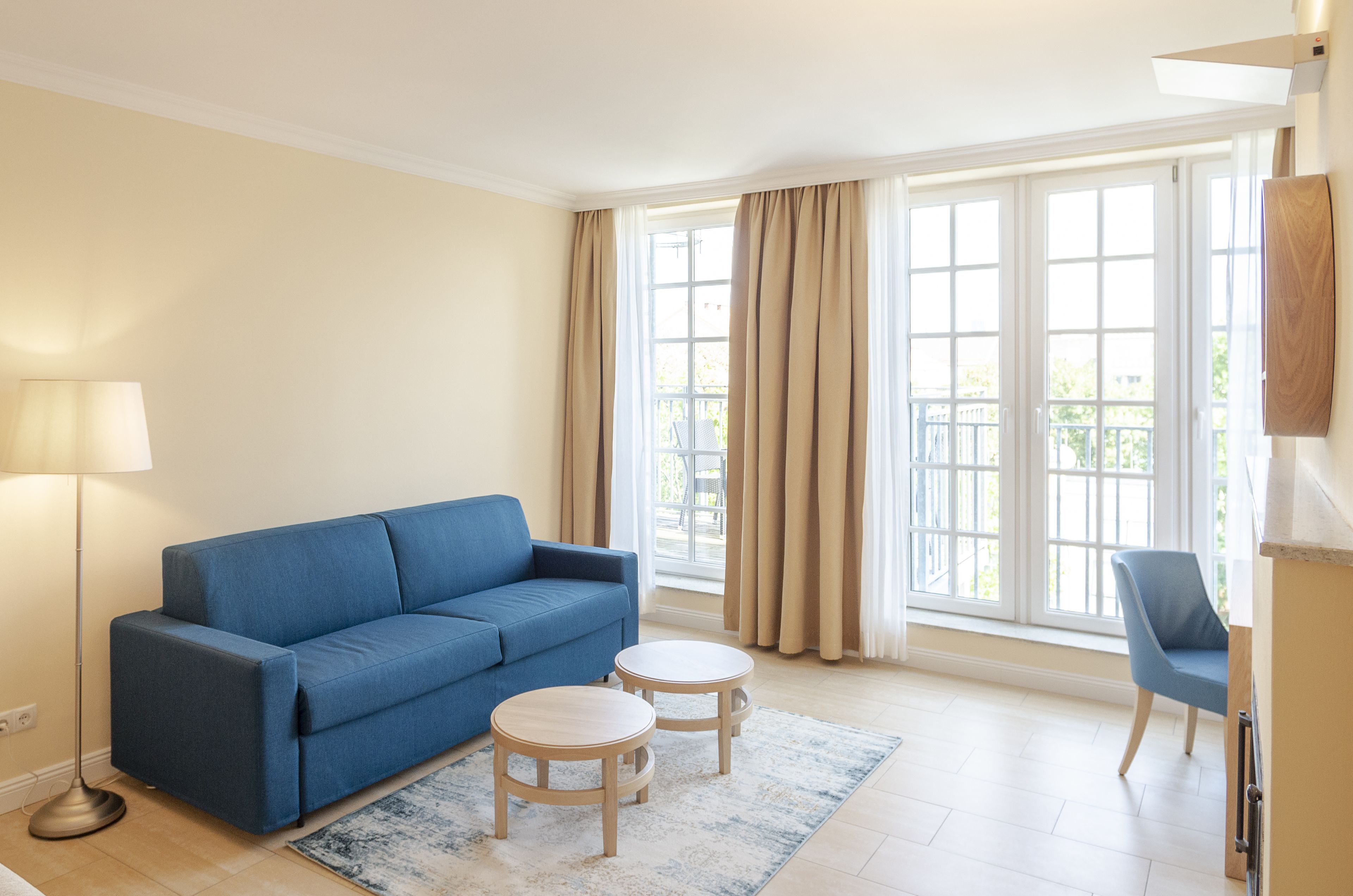 A modern living room with a blue sofa, two round wooden tables, a floor lamp, and large windows with curtains.