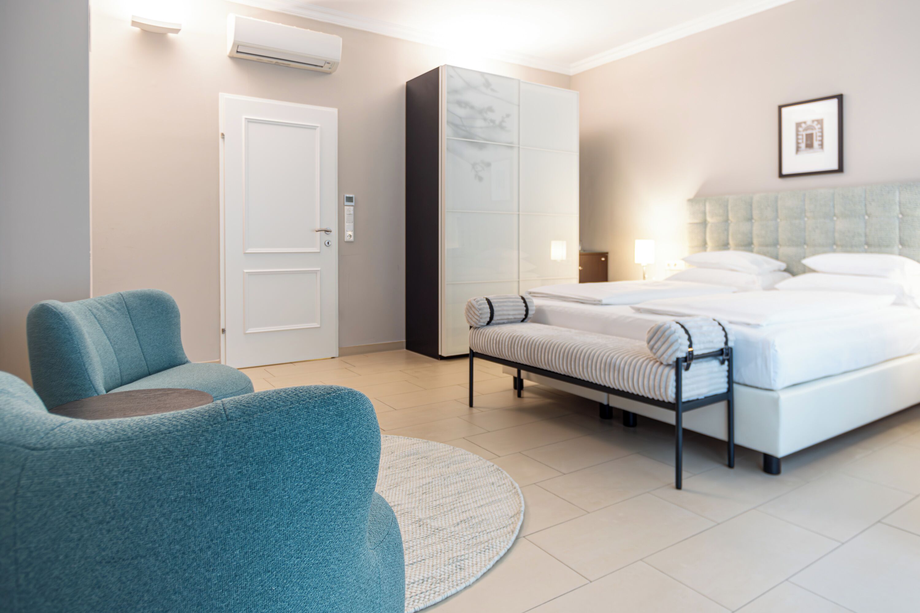 Una habitación de hotel moderna con una cama grande, una zona de estar con una silla azul, un armario y una pequeña mesa.