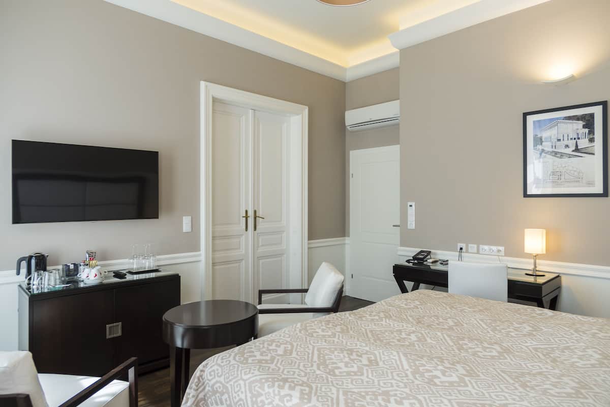 A hotel room with a bed, a desk, a chair, a small table, a minibar, a TV, and a framed picture on the wall.