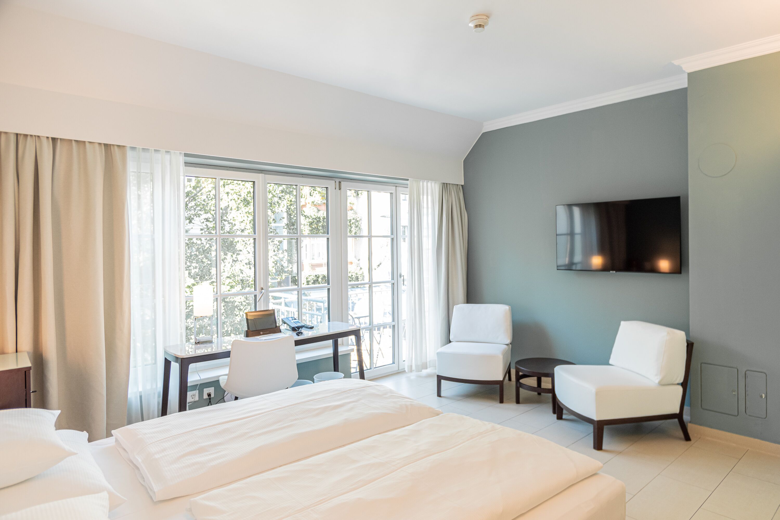 A hotel room with a large bed, two white armchairs, a desk with a chair, a flat-screen TV mounted on the wall, and a large window with curtains.
