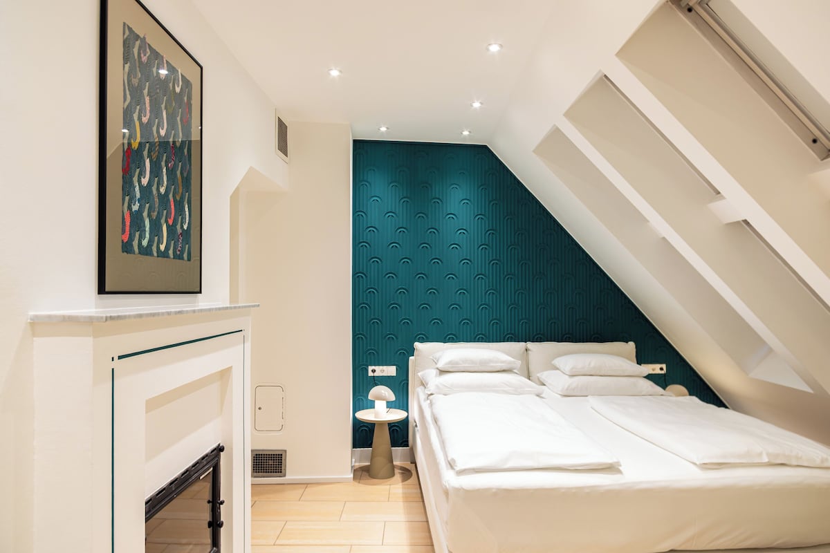 A modern bedroom with a sloped ceiling, a bed with white linens, a small bedside table, and a wall-mounted artwork.