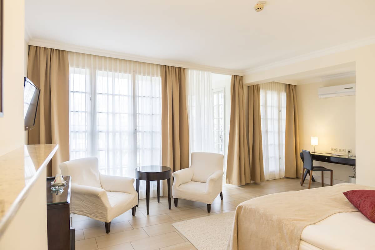 A hotel room with a bed, two armchairs, a small round table, a desk with a chair, and a TV.