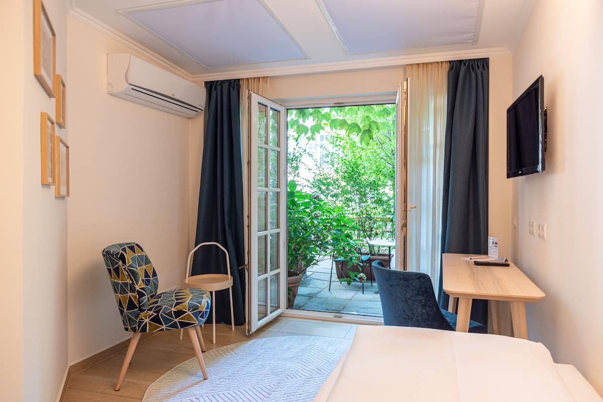 A hotel room with a bed, a desk, a chair, and a TV. There is a sliding glass door leading to an outdoor area with plants.