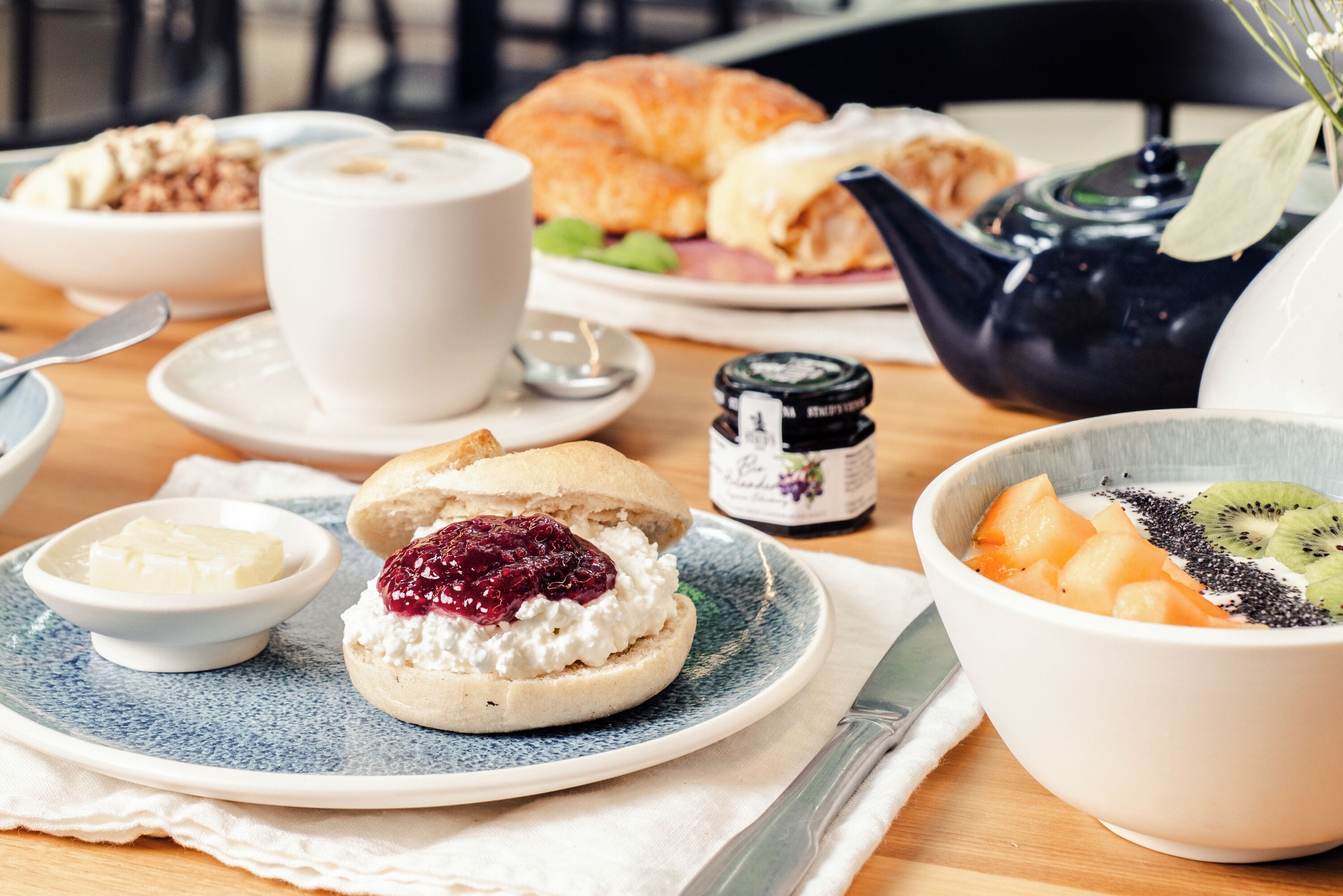 En frukostuppsättning med en croissant, en kopp kaffe, en skål med frukt och en tekanna på ett träbord.