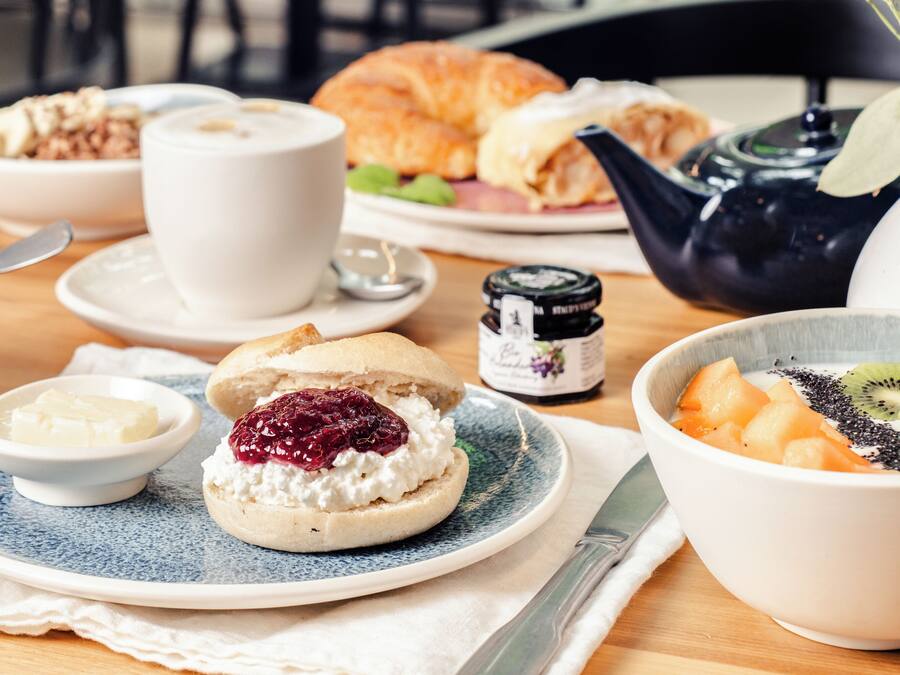 En frukostuppsättning med en croissant, en kopp kaffe, en skål med frukt och en tekanna på ett träbord.
