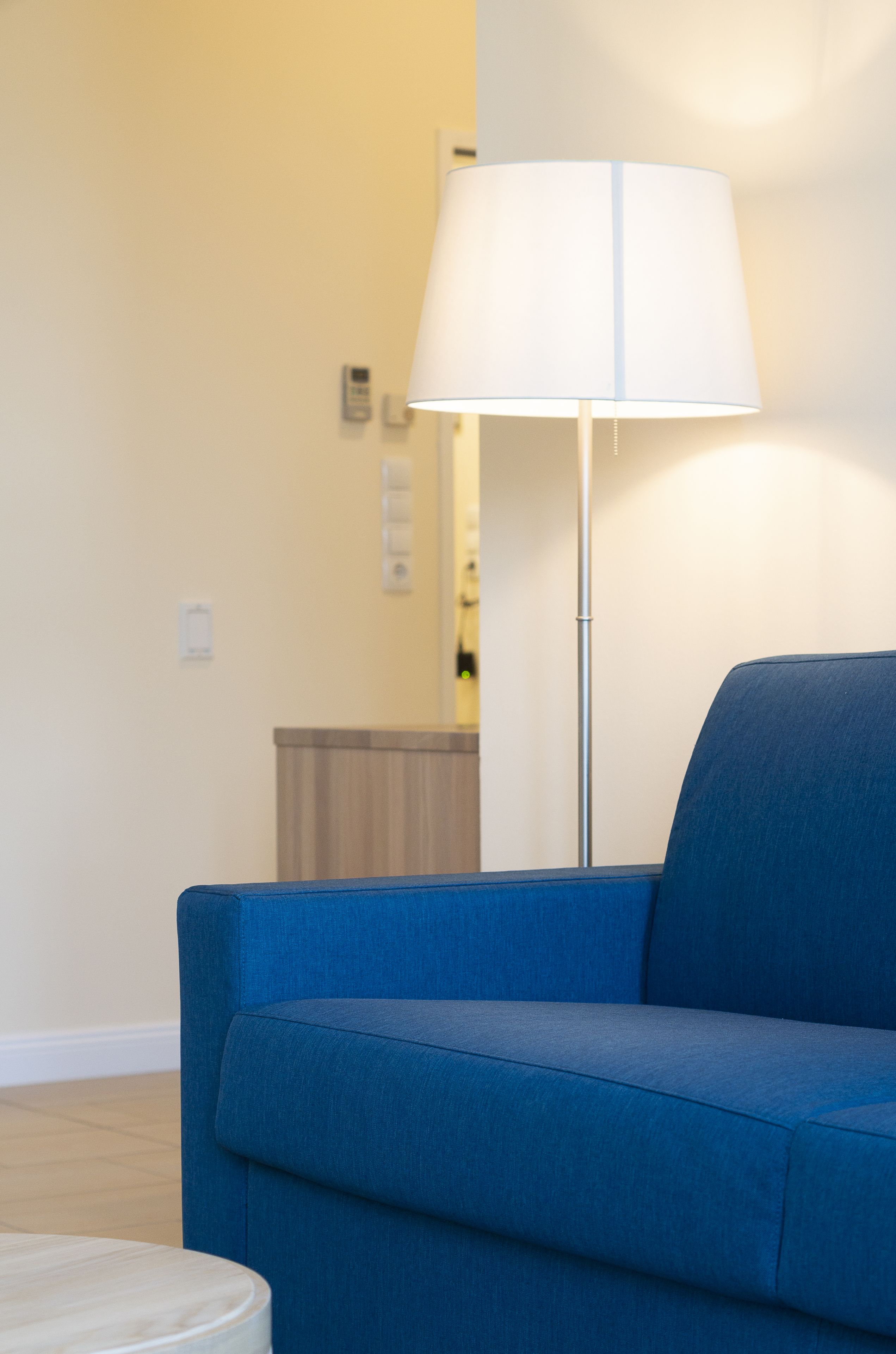 A blue sofa, a floor lamp, and a small round table in a room with plain walls and a light switch.