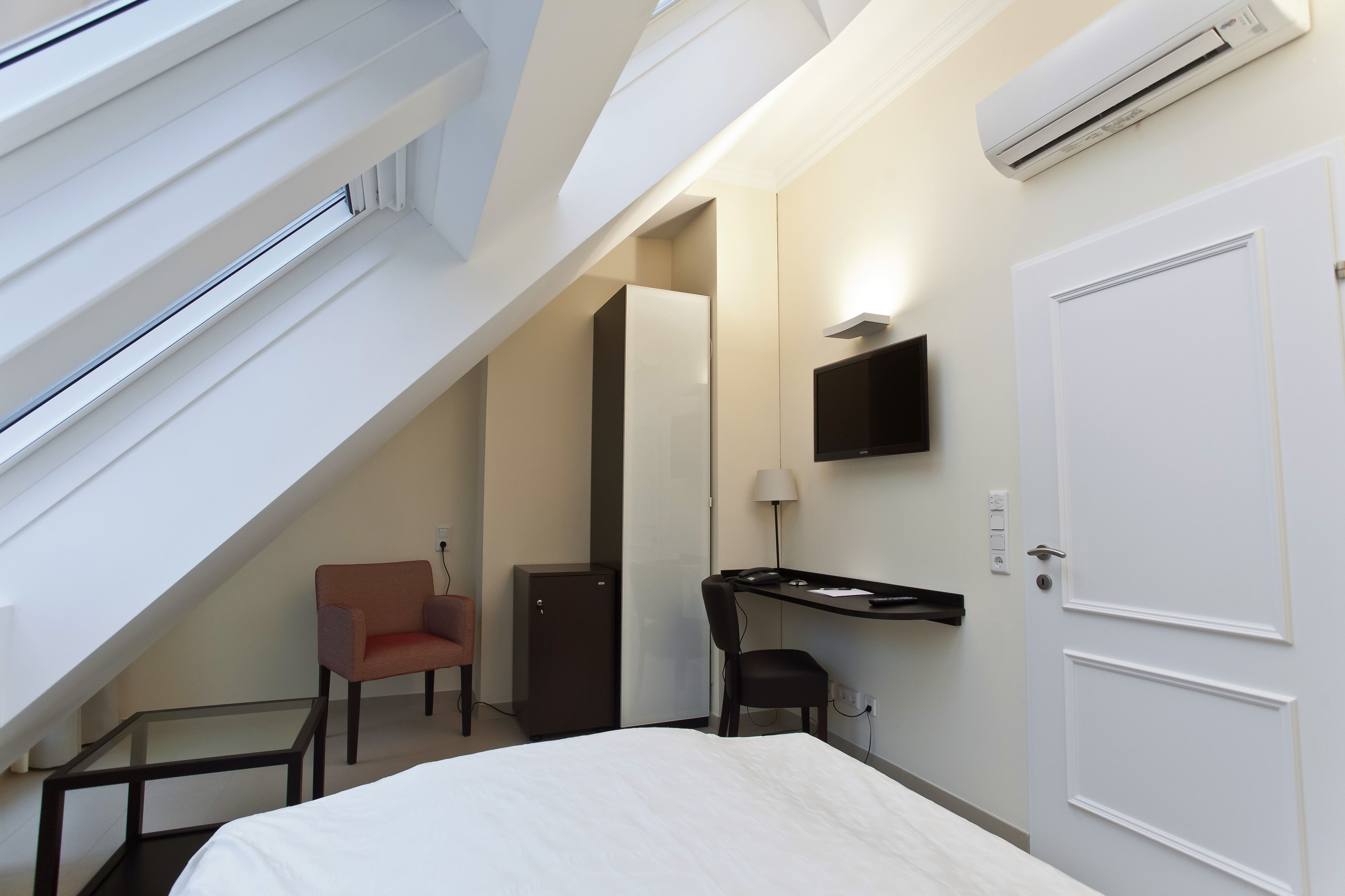Habitación de hotel con cama, escritorio, silla, mueble pequeño, televisor y aire acondicionado.