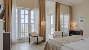 Una habitación de hotel con una cama, dos sillas, un escritorio, una lámpara y ventanas grandes con cortinas.