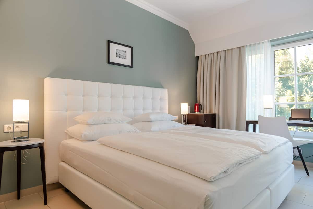 A hotel room with a large bed, a bedside table, a lamp, a framed picture on the wall, and a window with curtains.
