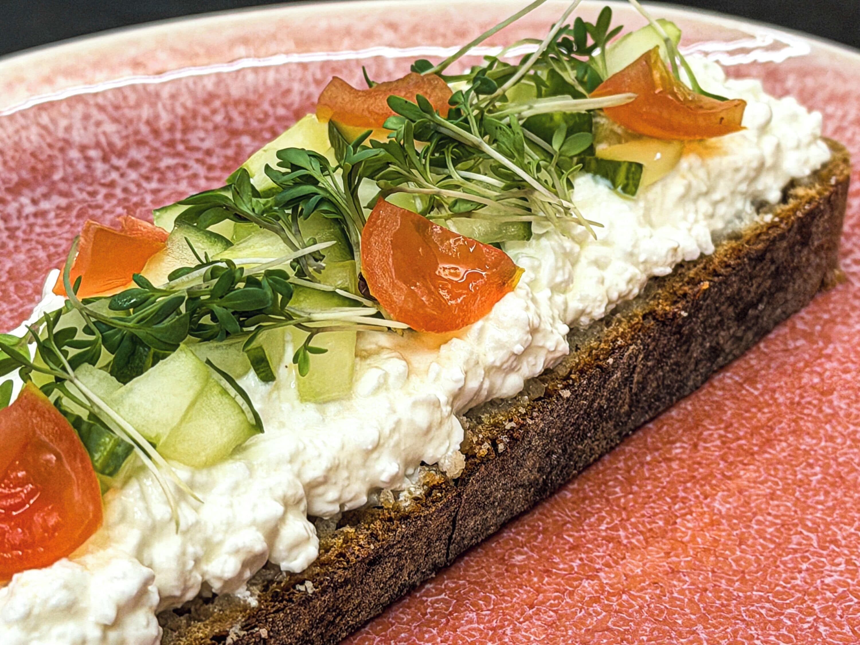 A slice of bread with cream cheese, cucumber, tomato, and greens.