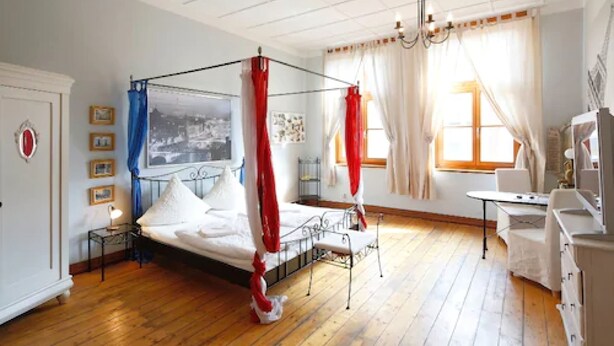 Een slaapkamer met een hemelbed, houten vloeren, een nachtkastje, een stoel en een raam met gordijnen.