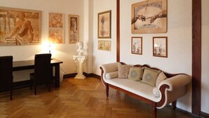 Een kamer met een houten visgraatvloer, een witte bank met gekleurde kussens, een witte bijzettafel en een bureau met lamp.