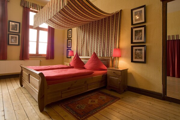 Una cama doble con ropa de cama roja, un cabecero de madera, una mesita de noche con una lámpara, una alfombra estampada y una ventana con cortinas.