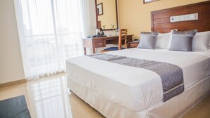 A hotel room with a large bed, a desk, and a chair. There is a window with curtains, a mirror on the wall, and a thermostat.