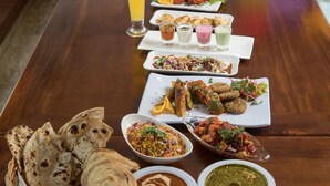 A long wooden table with various dishes, including flatbreads, curries, and a tall glass of a yellow beverage.