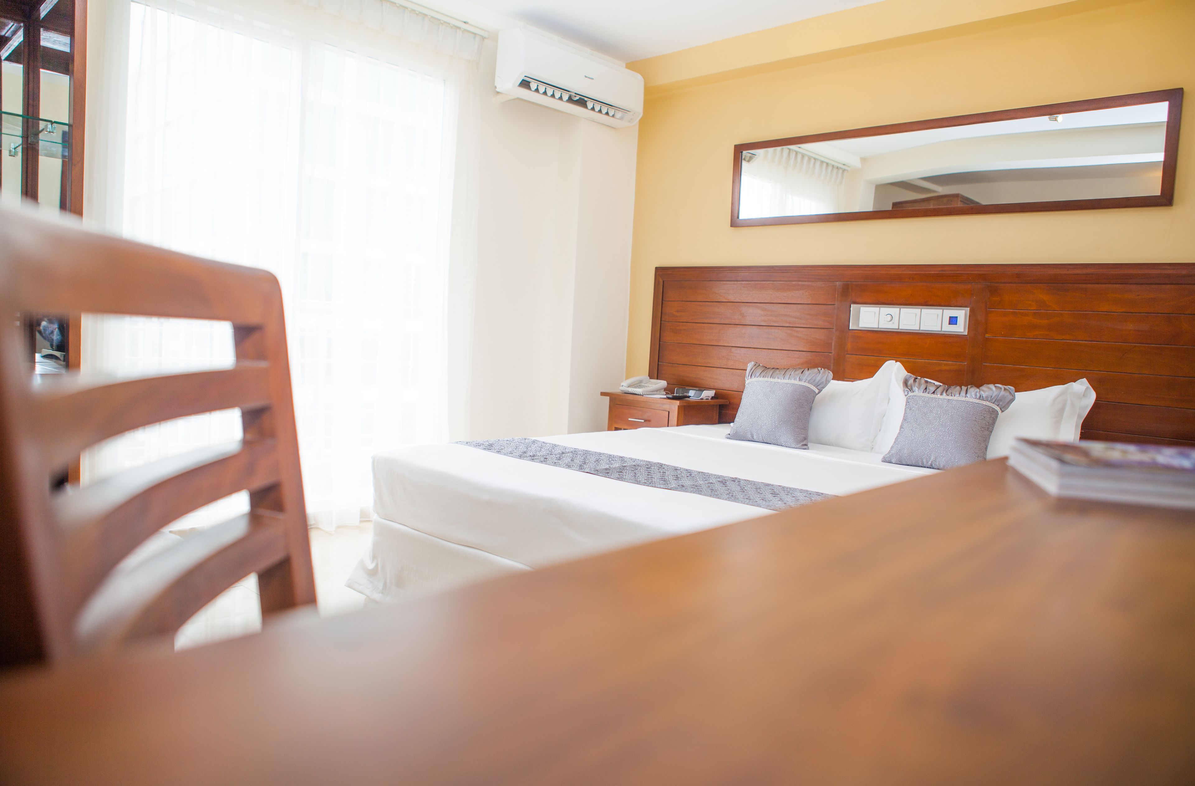 Een hotelkamer met een houten hoofdeinde, een nachtkastje met een lamp, een grote spiegel en een raam met gordijnen.