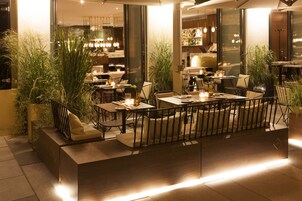 An outdoor dining area with tables and chairs, surrounded by potted plants and illuminated by hanging lights.
