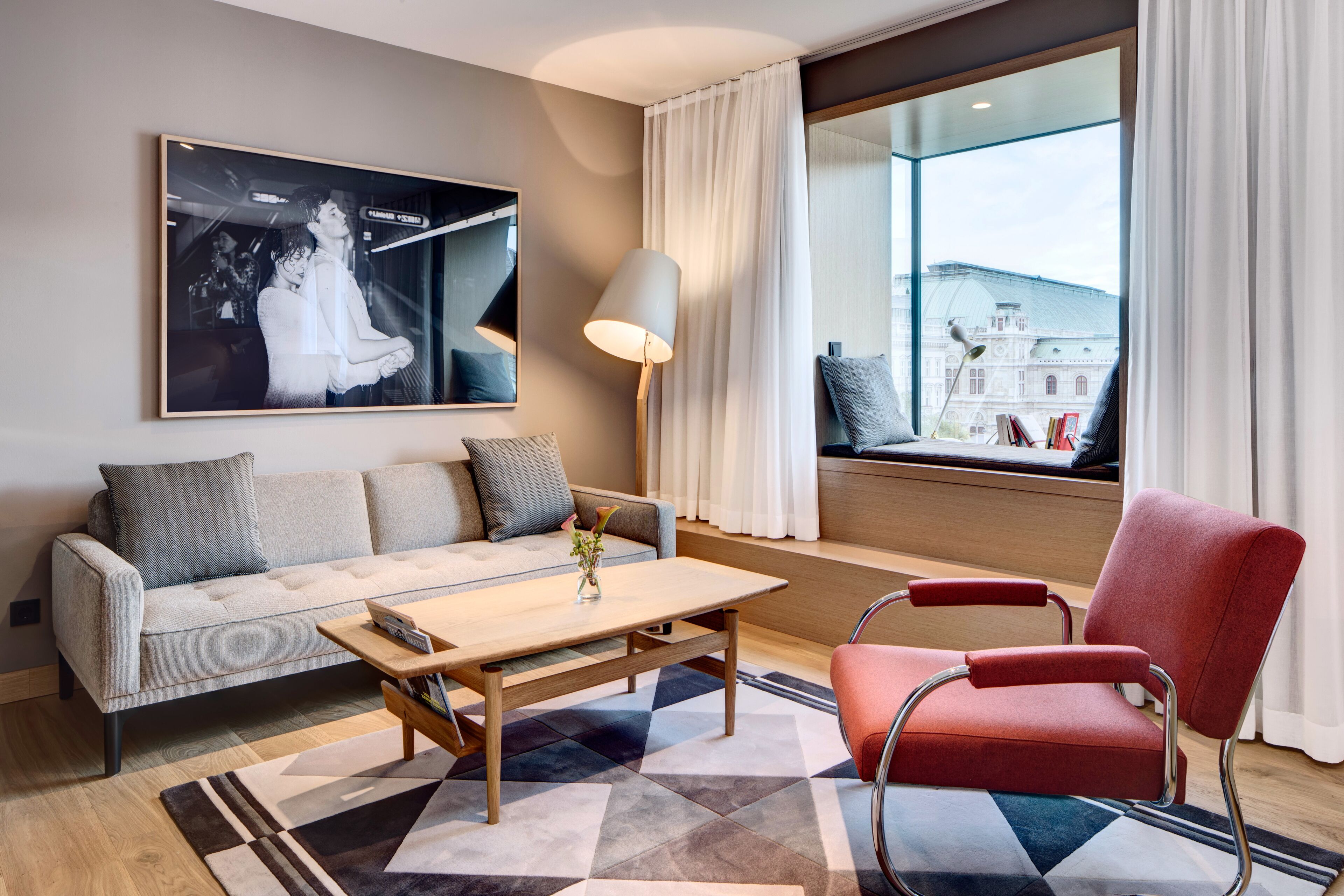 Ein modernes Hotelzimmer mit einer Couch, einem roten Sessel, einem Holztisch und einem großen Fenster mit Blick auf ein Gebäude.