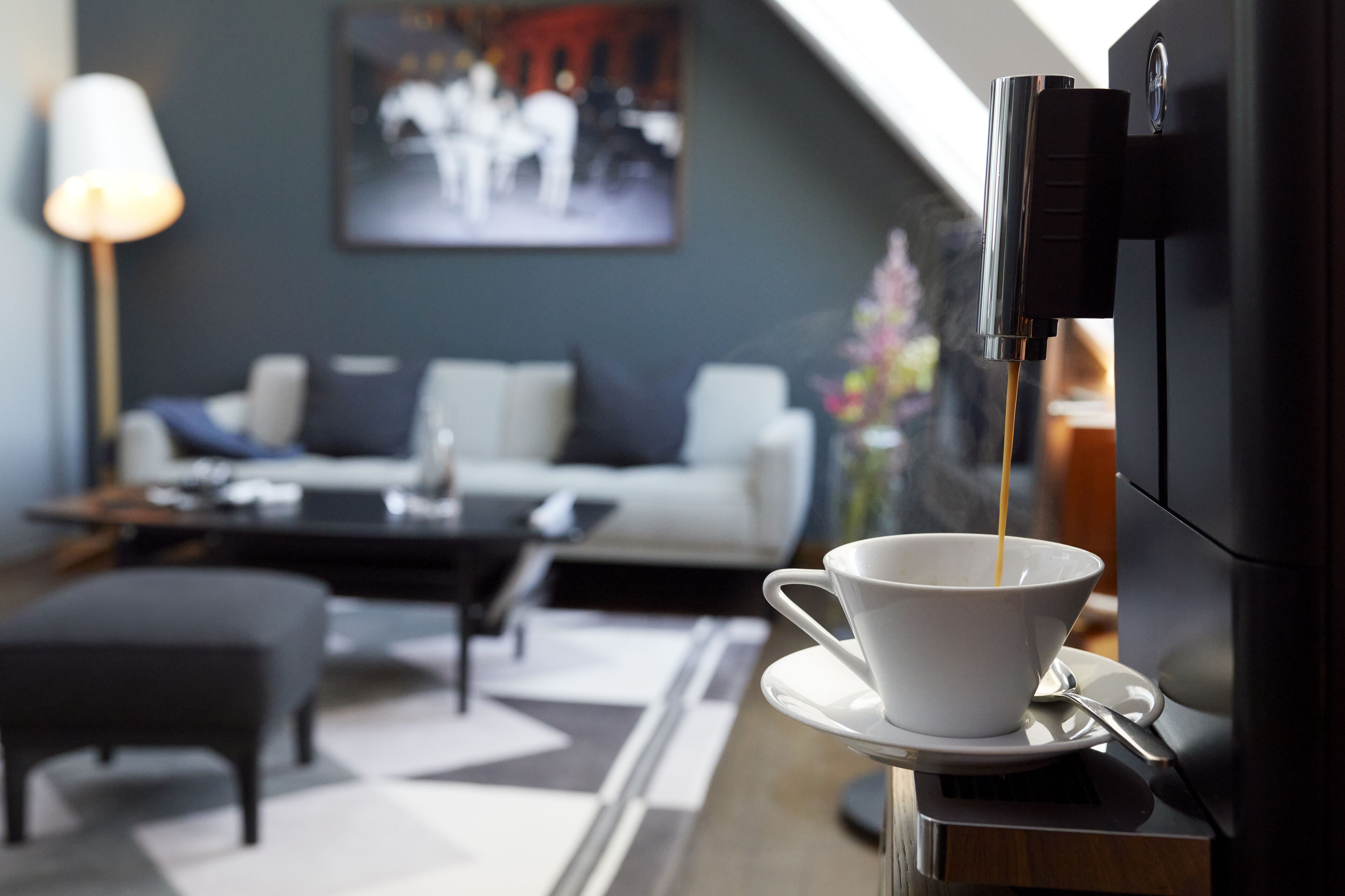 Ein modernes Wohnzimmer mit Kaffeemaschine, einer weißen Tasse auf Untertasse und einem Sofa mit Kissen.