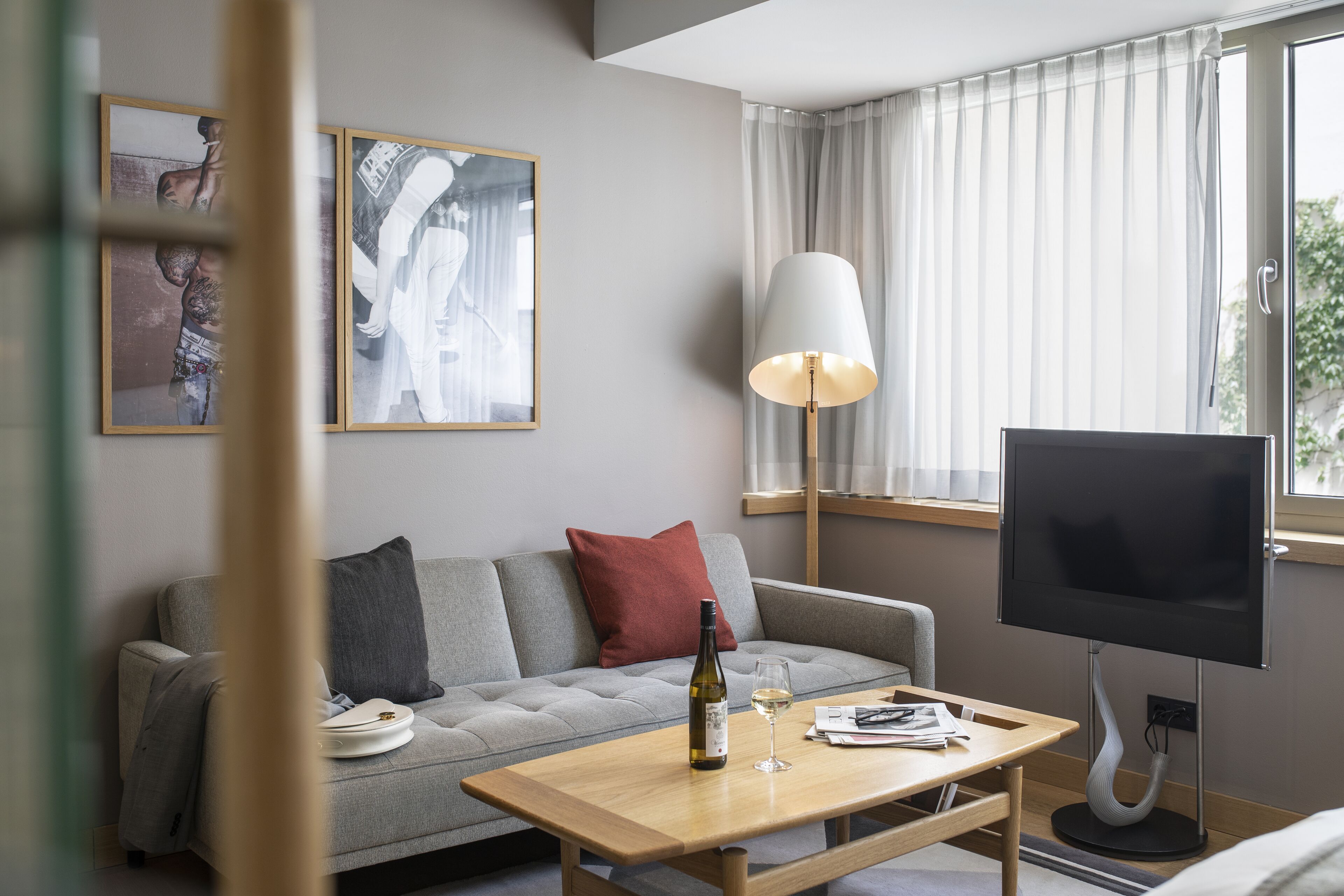 A modern living room with a grey sofa, a wooden coffee table with a bottle and glass, a flat-screen TV, and a floor lamp.