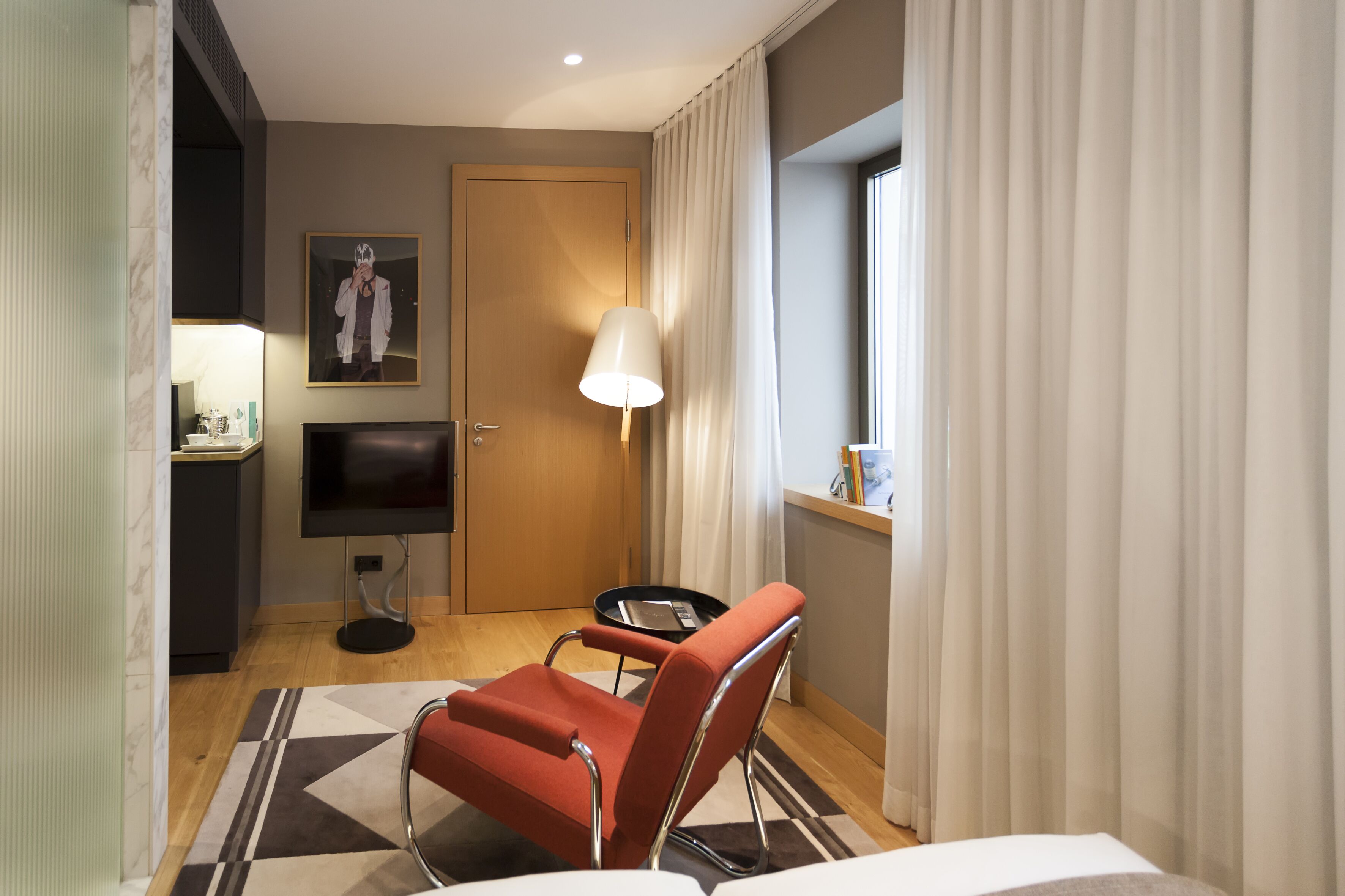 Una habitación de hotel con cama, una silla roja, televisión, lámpara y ventana con cortinas.