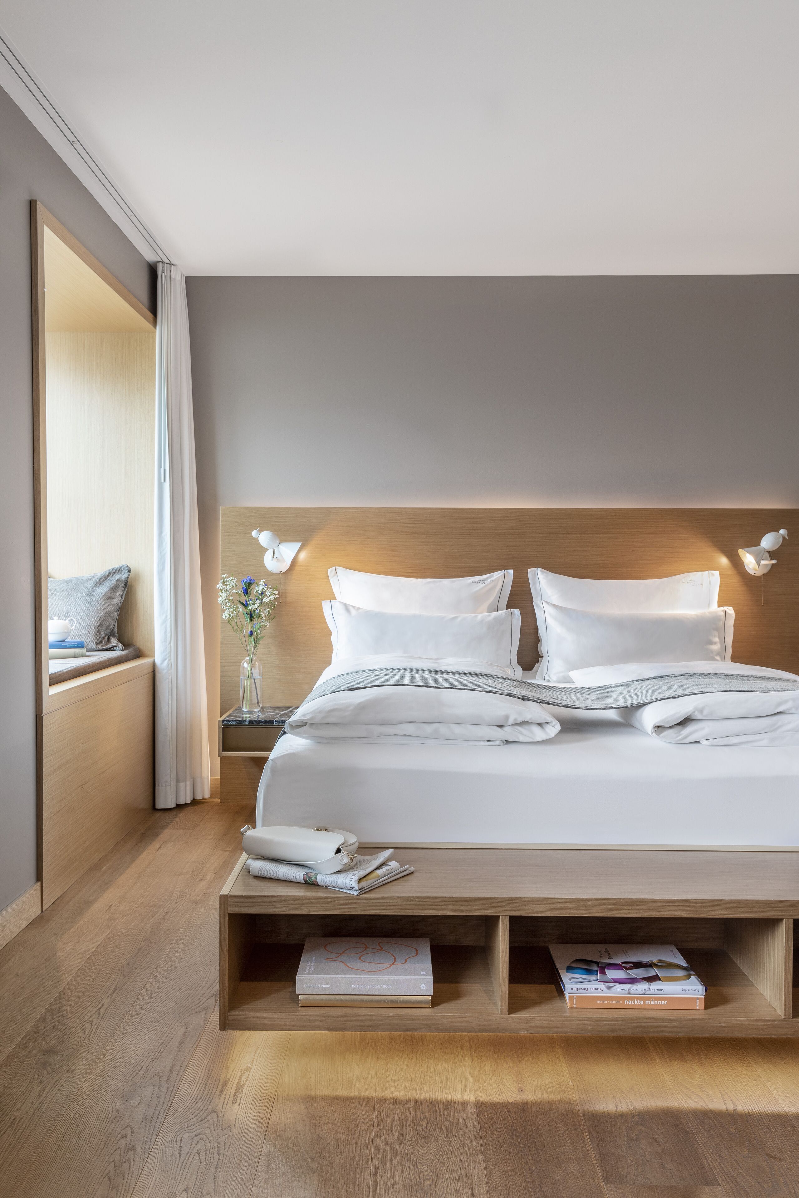 A modern hotel room with a wooden headboard, white bedding, a bedside table with a vase of flowers, and a built-in bookshelf.