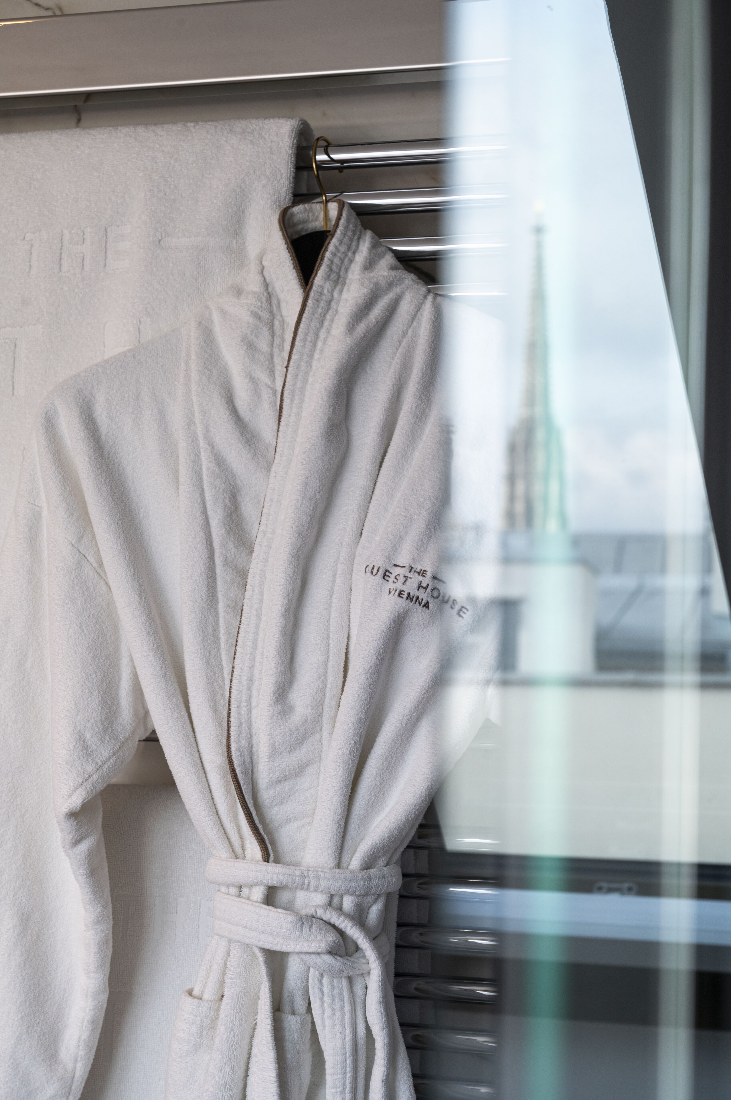 A white bathrobe hanging on a metal rack with a window view of a building.