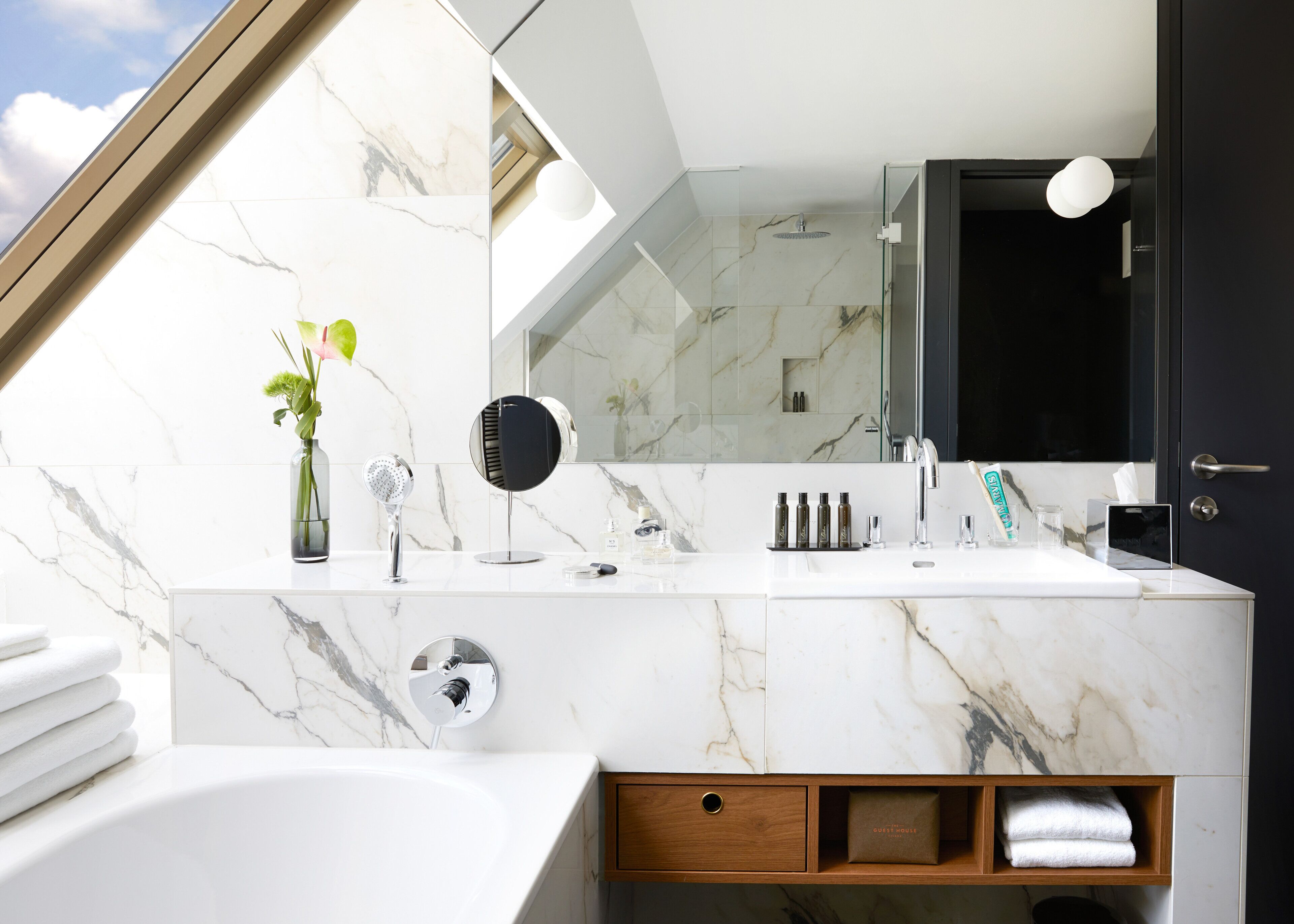 A modern bathroom with a marble vanity, a large mirror, a shower, and a wooden cabinet with towels.