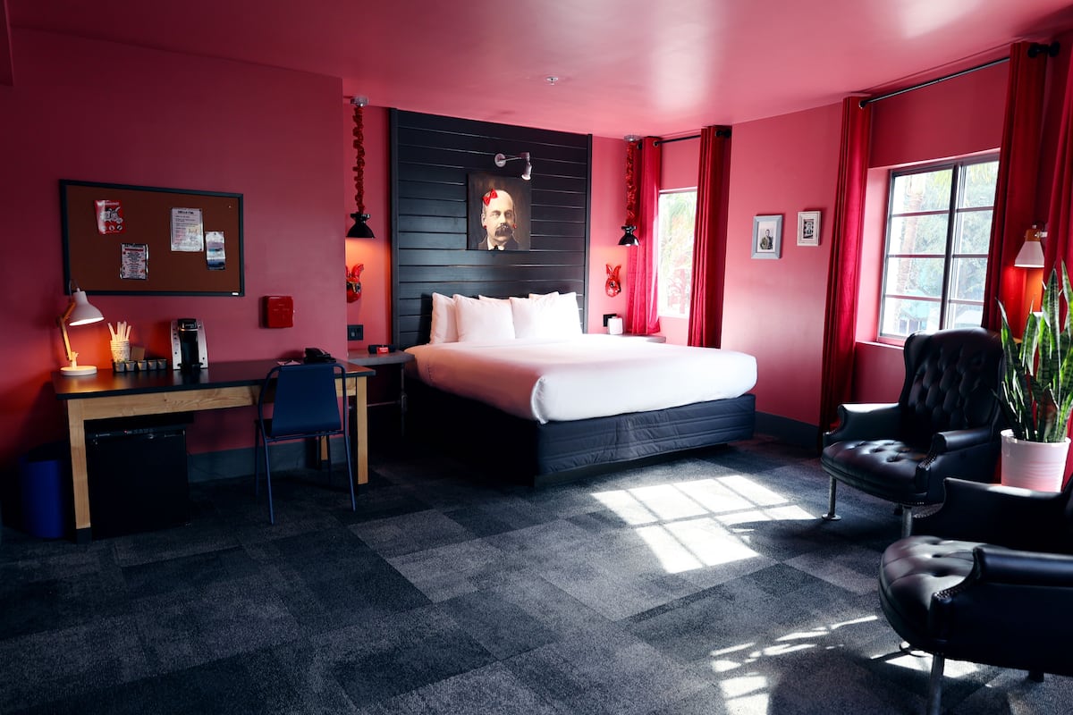 Une chambre d’hôtel moderne, dotée d’un grand lit, d’un bureau et d’une chaise. On y trouve également un tableau d’affichage avec des notes et une fenêtre avec des rideaux.