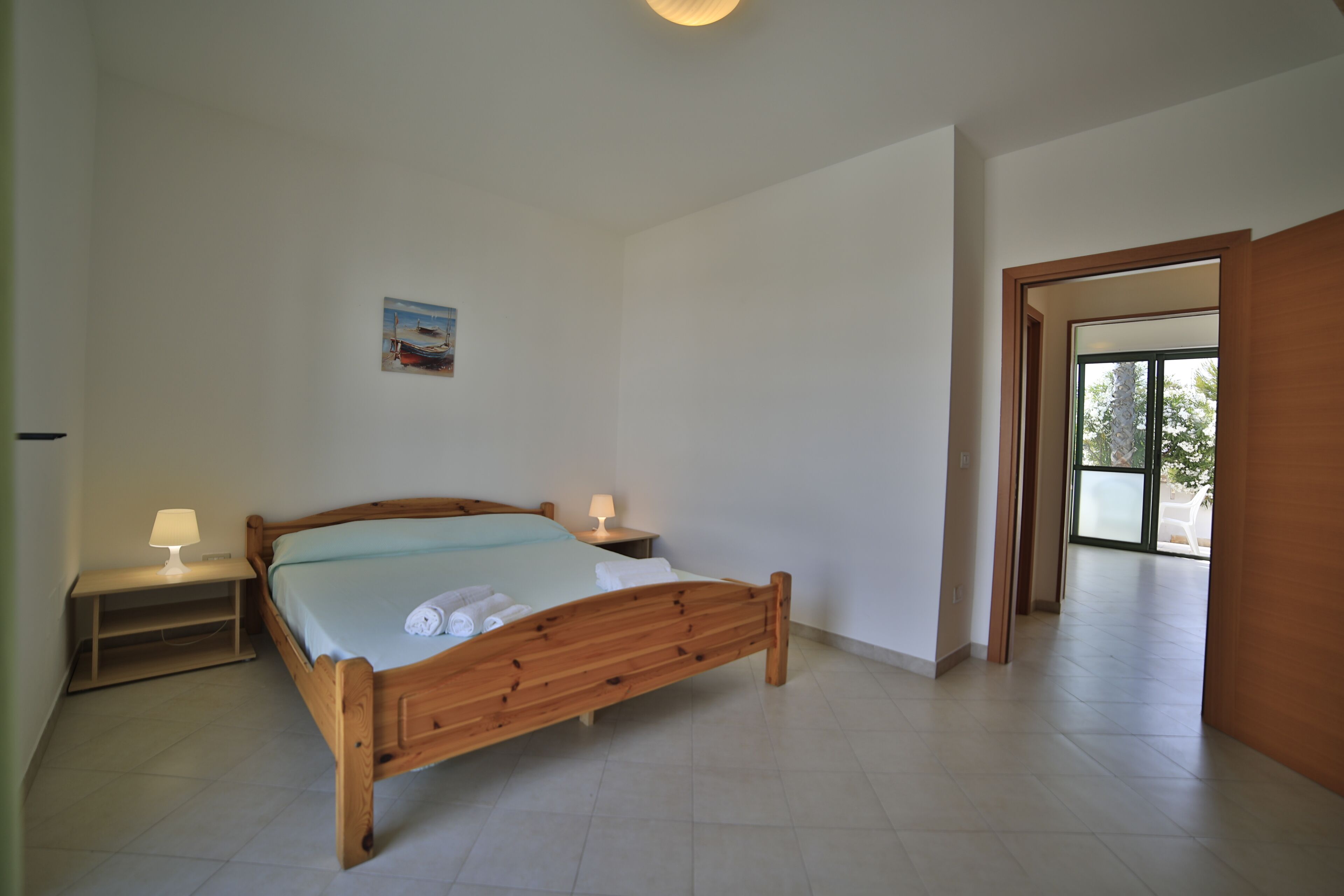 Een slaapkamer met een houten bed, twee nachtkastjes met lampen, een aan de muur hangend schilderij en een deur die naar een andere kamer leidt.
