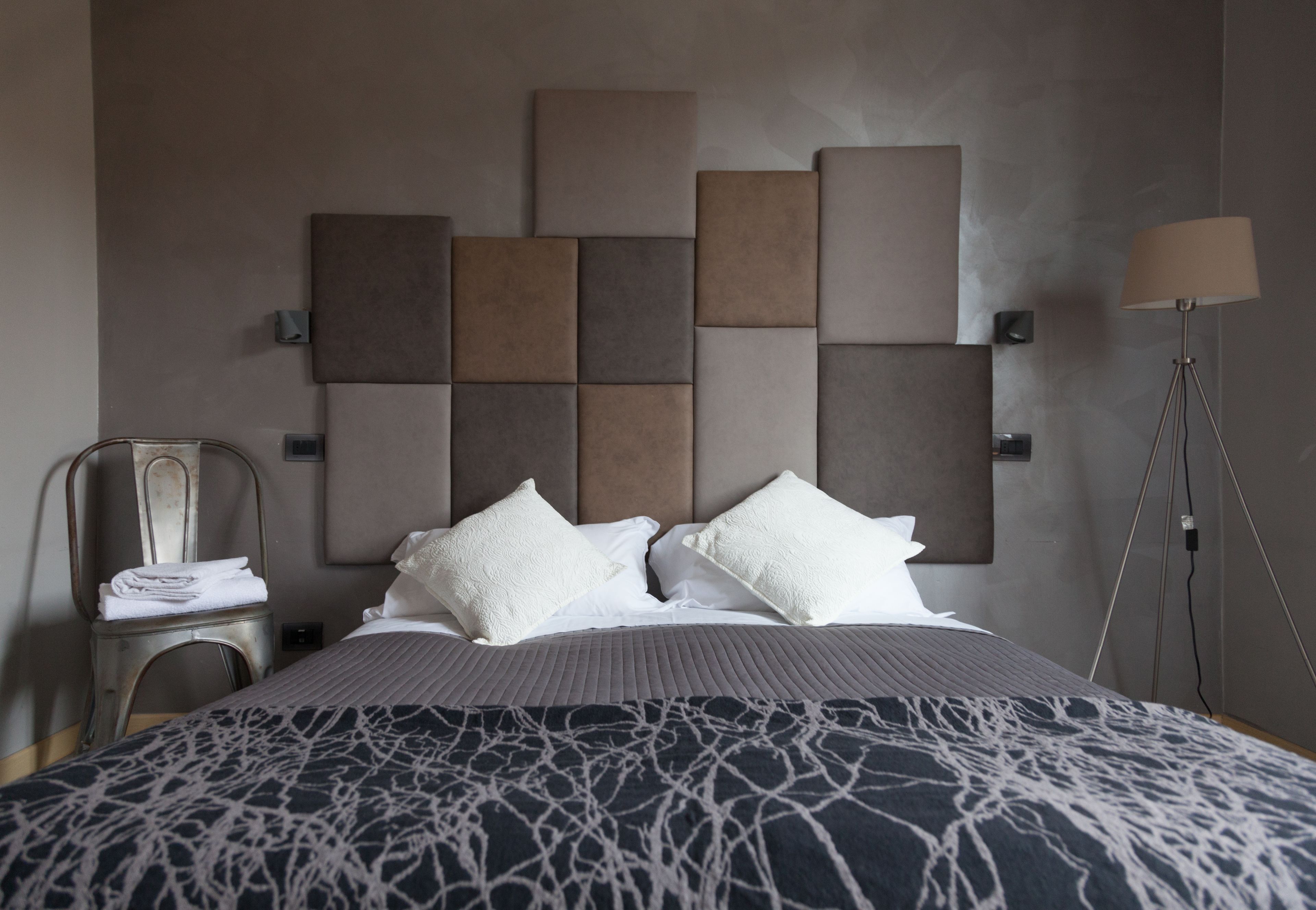 A modern hotel room with a large bed, a headboard with geometric panels, a bedside table with a lamp, and a chair with a towel.