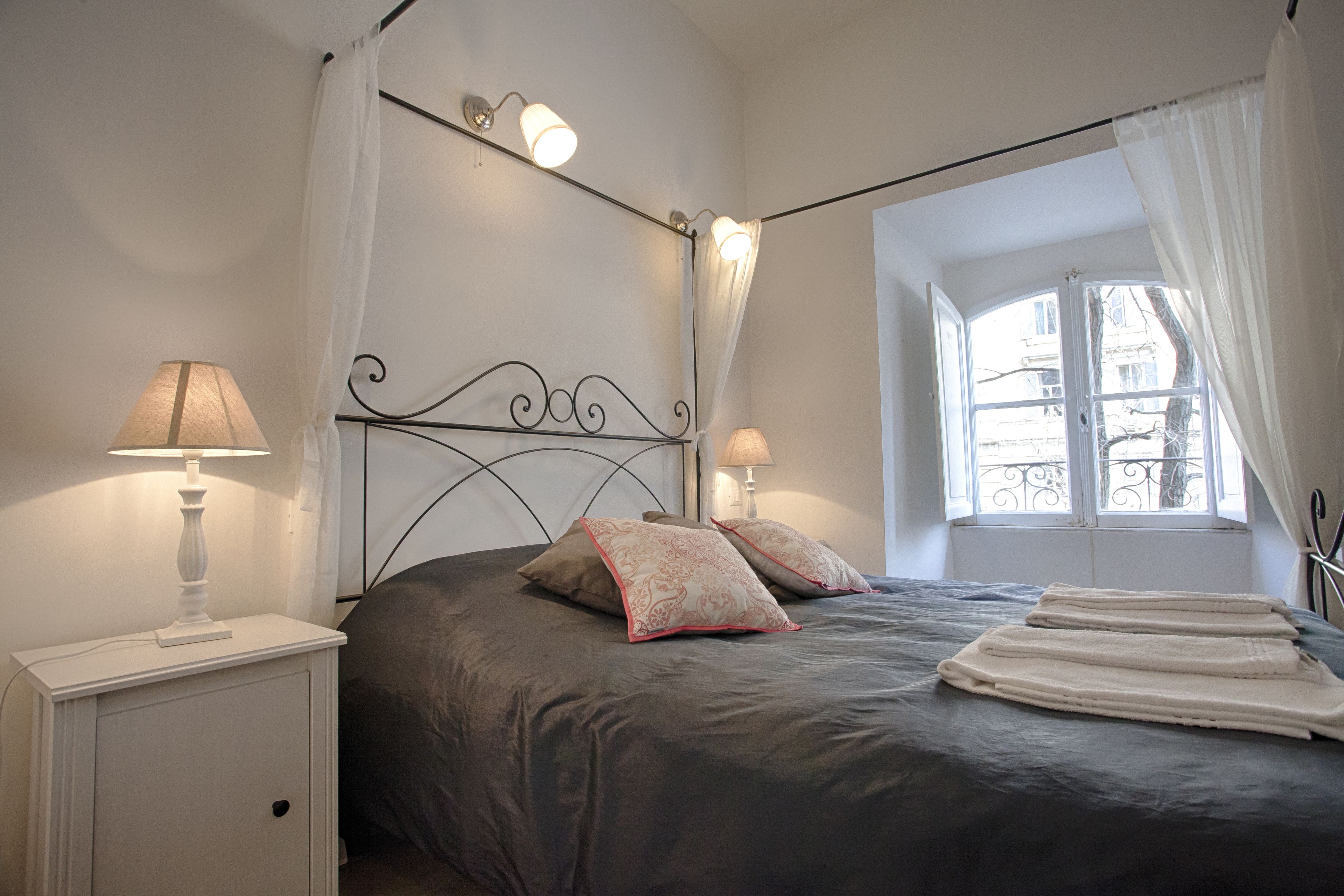 A bedroom with a bed, two bedside lamps, a window with curtains, and a nightstand with a lamp.